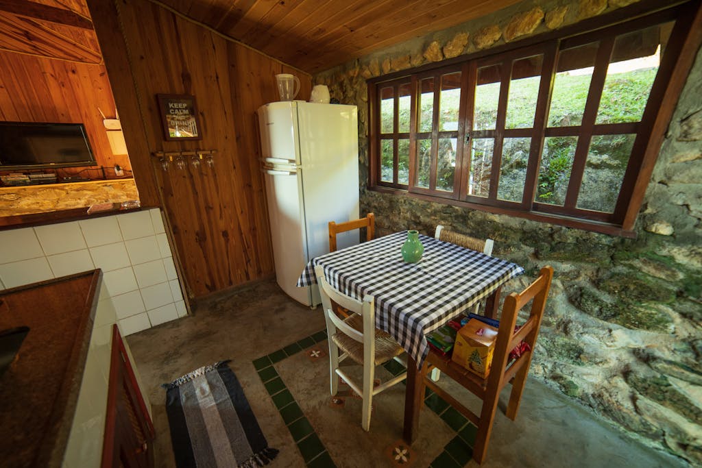 Chairs at table placed near refrigerator at stone wall with window in rural style kitchen with decoration on wooden wall